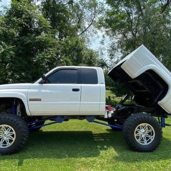 A white lifted pickup truck equipped with the PIERCE Factory Bed Dump Kit has its bed tilted up, parked on grass with trees in the background.