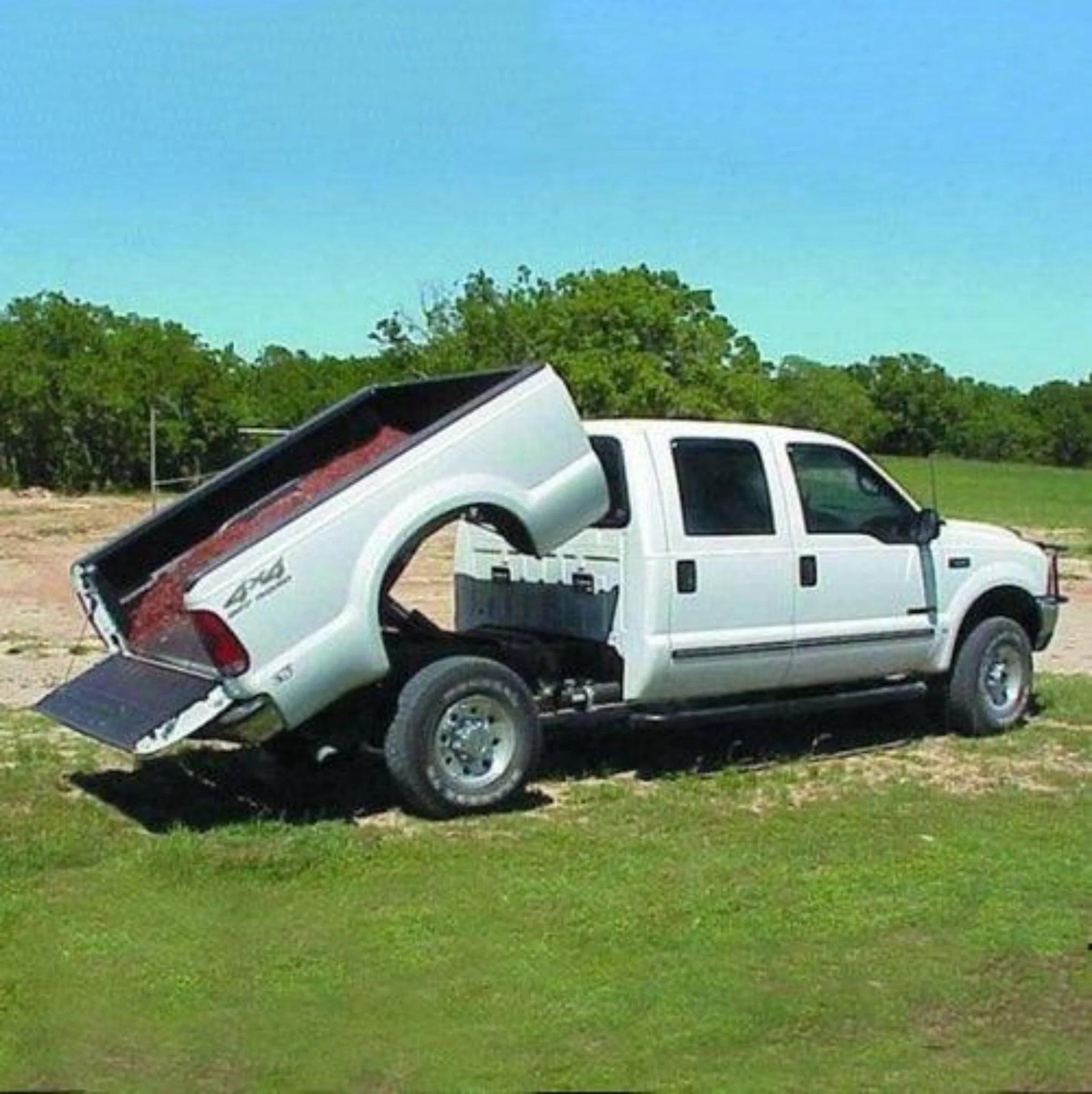 A white pickup with a PIERCE Factory Bed Dump Kit has its bed unusually tilted upward, separated from the cab, and is parked on grass.