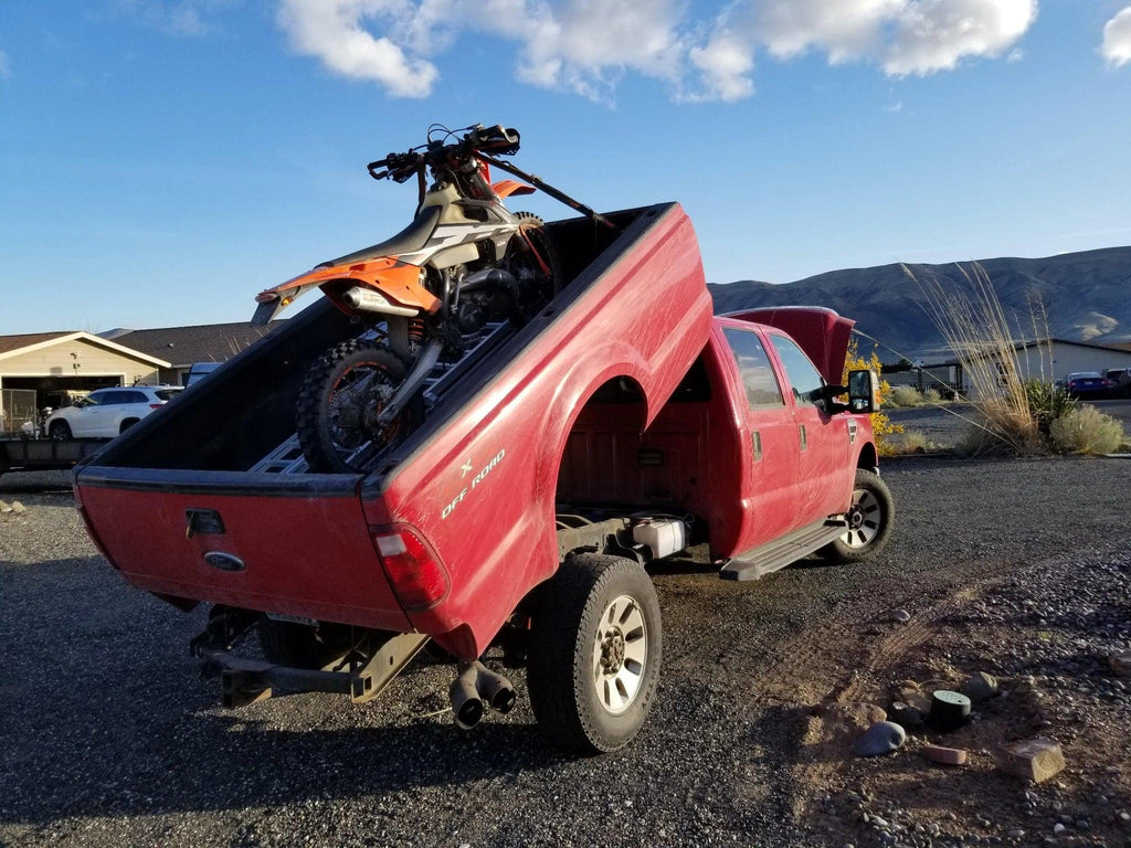 A red pickup truck equipped with a PIERCE Factory Bed Dump Kit tilts its bed up to hold a dirt bike, parked on gravel near mountains.