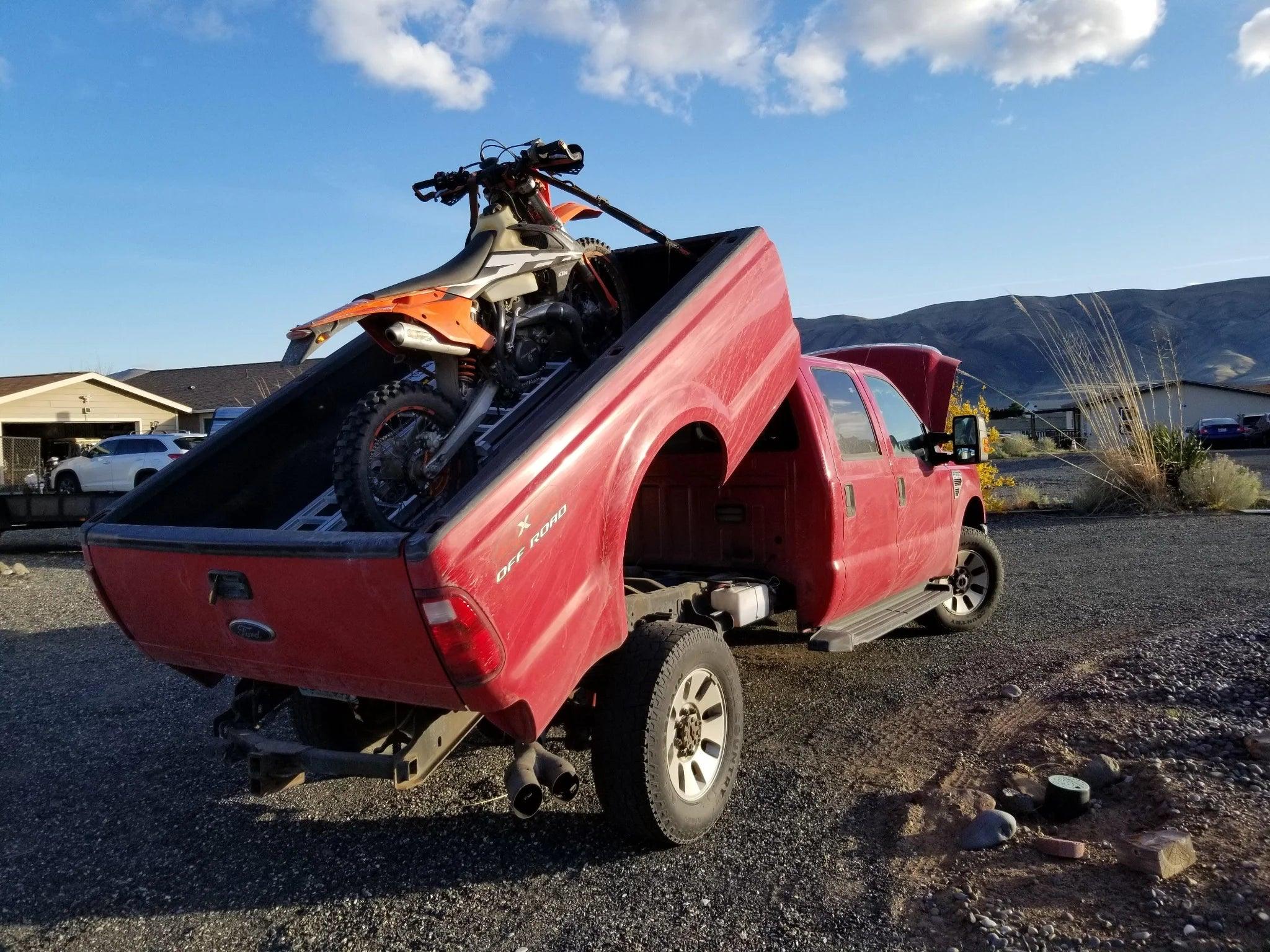 A red pickup truck equipped with a PIERCE Factory Bed Dump Kit tilts its bed up to hold a dirt bike, parked on gravel near mountains.