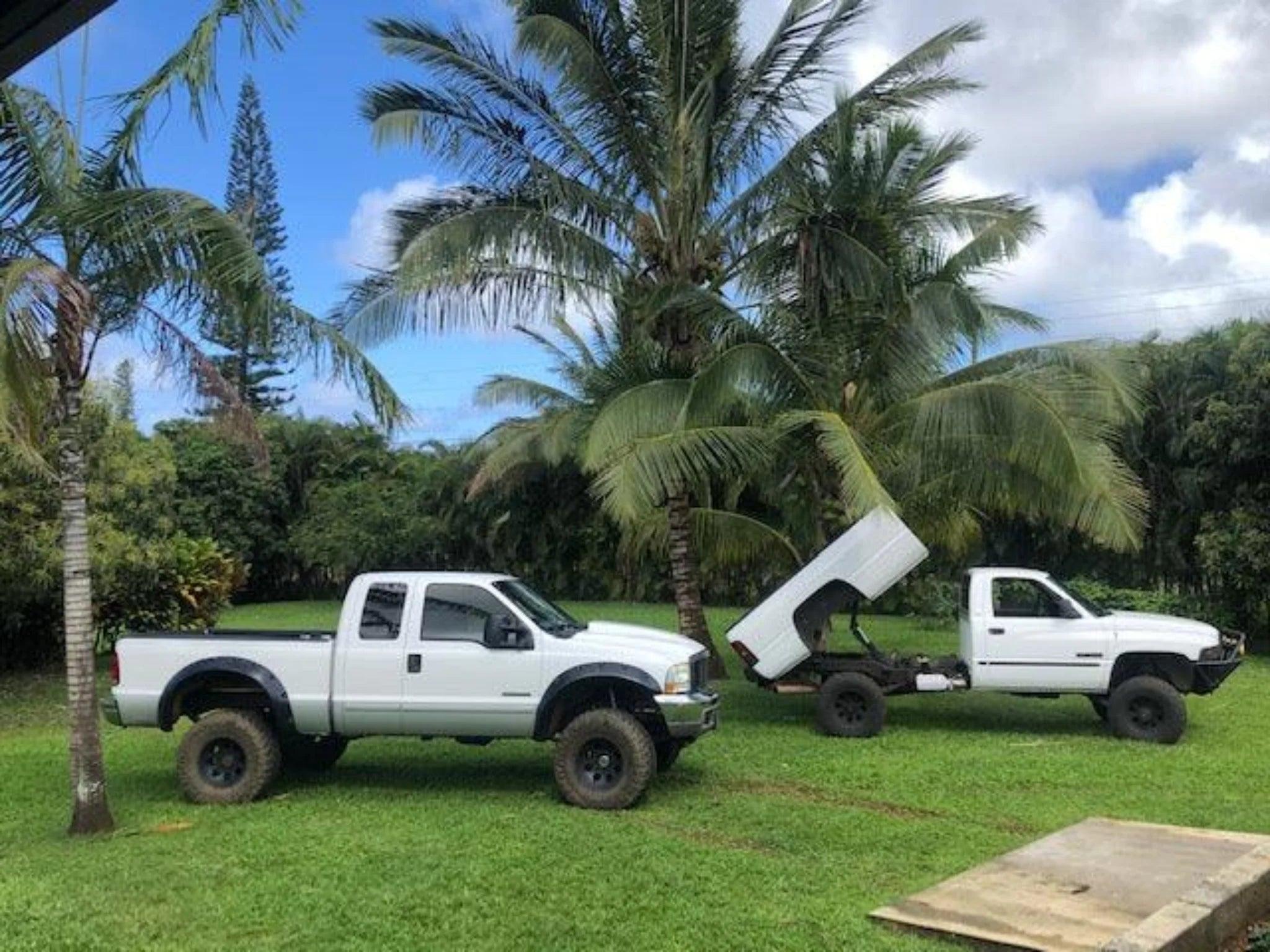 Two white pickup trucks on grass by palm trees; one has a raised bed with exposed frame, highlighting its PIERCE Factory Bed Dump Kit hydraulic system.