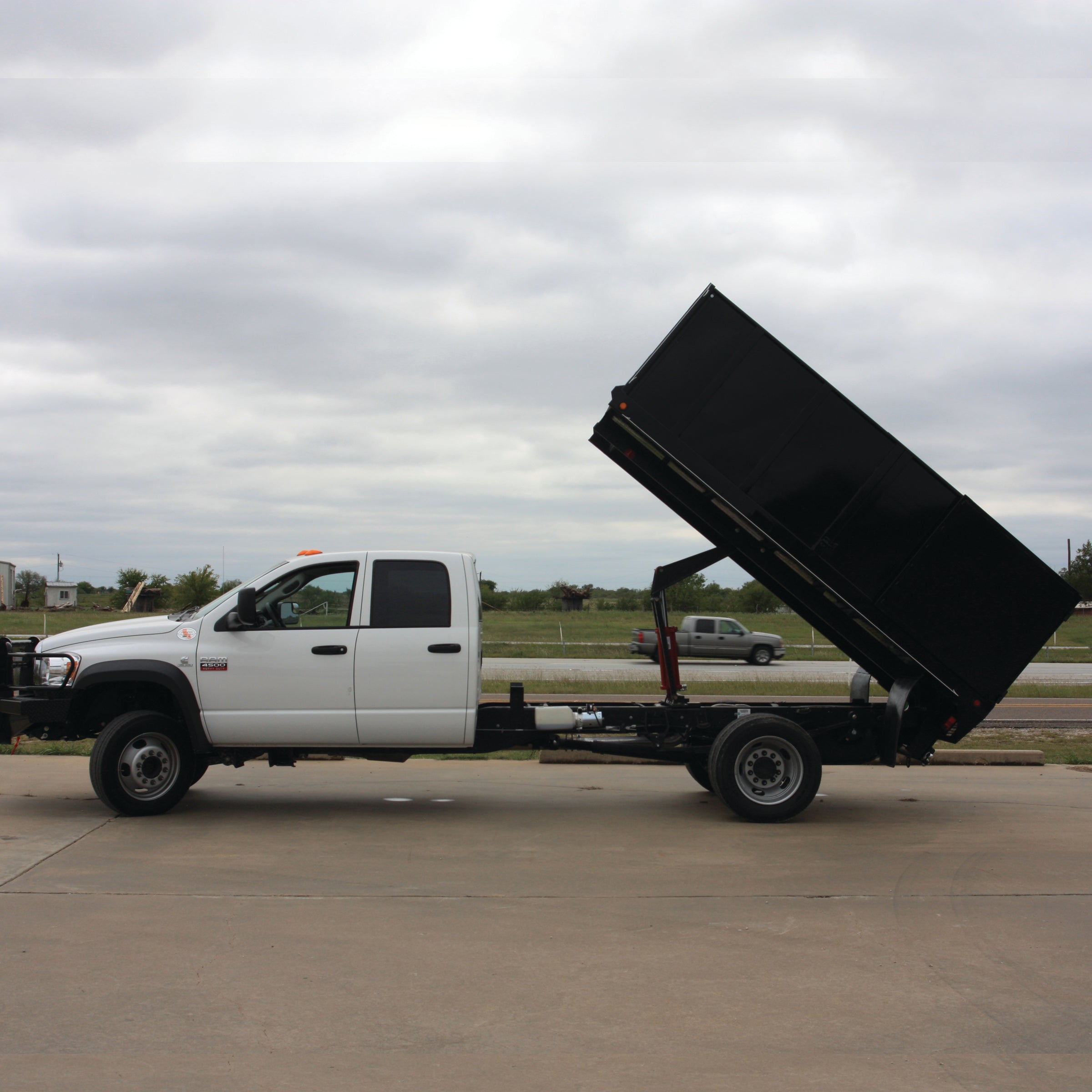A white truck with a PIERCE Heavy Duty Flat Bed Dump Kit, featuring a 12 Volt Pump, is parked on concrete under an overcast sky, its bed impressively raised.