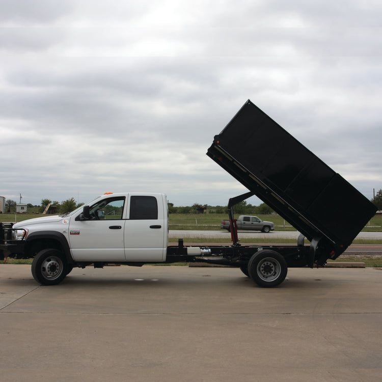 A white truck with a PIERCE Heavy Duty Flat Bed Dump Kit, featuring a 12 Volt Pump, is parked on concrete under an overcast sky, its bed impressively raised.