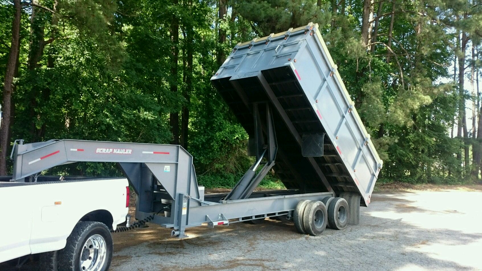 A pickup truck in a tranquil wooded area lifts a dump trailer with the PIERCE Heavy Duty Flat Bed Dump Kit - 12 Volt Pump, raising it at a steep angle.