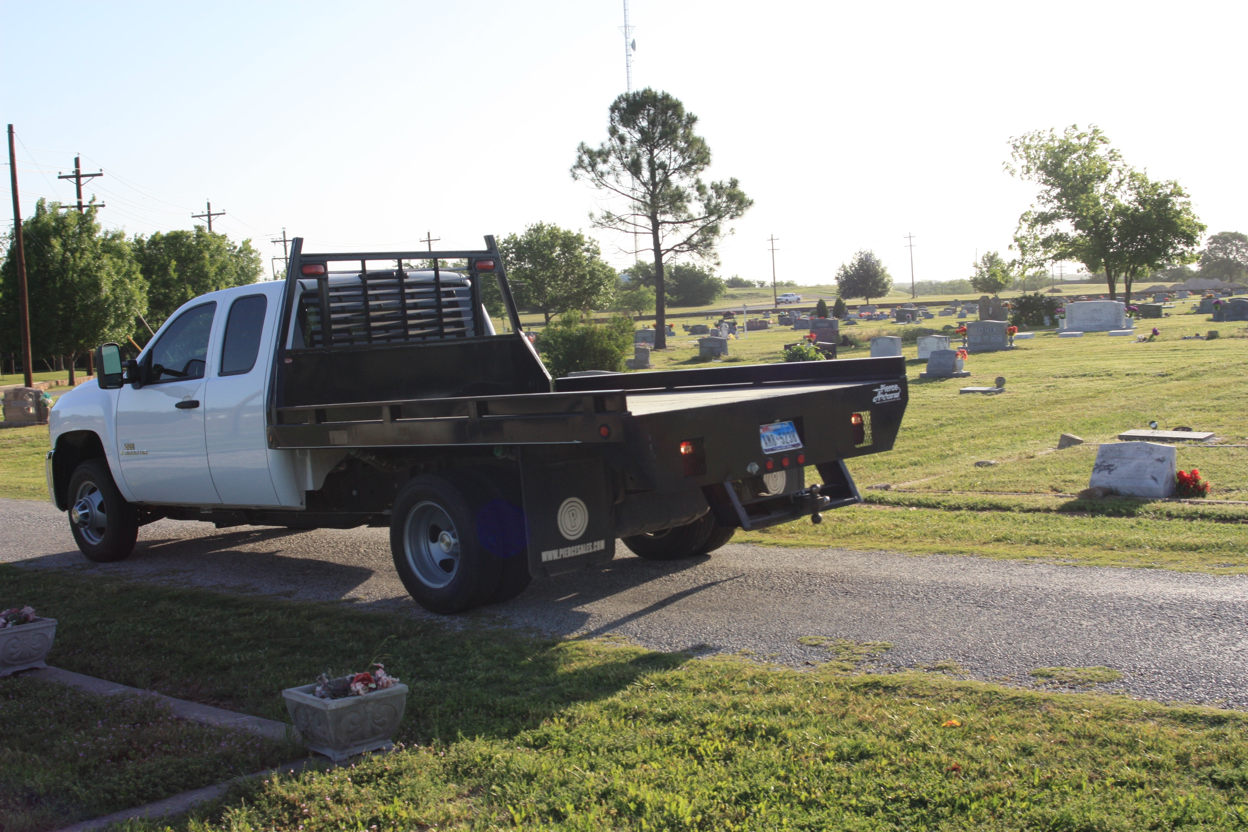 A white flatbed truck with a PIERCE Heavy Duty Flat Bed Dump Kit featuring a 12 Volt Pump is parked on a cemetery path, surrounded by gravestones and trees.