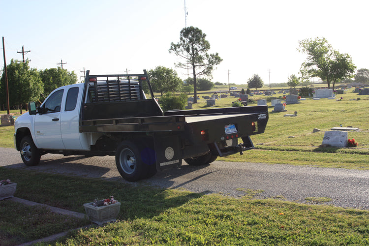 A white flatbed truck with a PIERCE Heavy Duty Flat Bed Dump Kit featuring a 12 Volt Pump is parked on a cemetery path, surrounded by gravestones and trees.