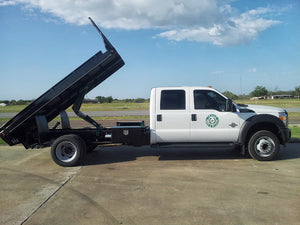 A white truck with a raised black dump bed, likely fitted with a PIERCE Heavy Duty Flat Bed Dump Kit - 12 Volt Pump, is parked on a concrete surface under a partly cloudy sky.