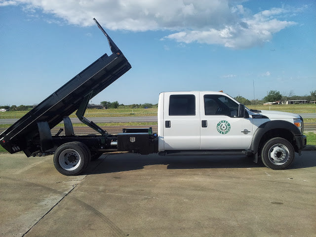 A white truck with a raised black dump bed, likely fitted with a PIERCE Heavy Duty Flat Bed Dump Kit - 12 Volt Pump, is parked on a concrete surface under a partly cloudy sky.