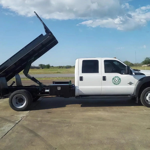 PIERCE Medium Duty Flat Bed Dump Kit installed on a white crew cab dump truck with raised black bed, parked on concrete beside a grassy field under partly cloudy skies.