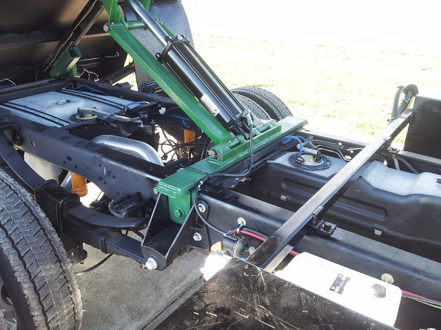 The side view of the truck bed lift mechanism shows a green metal arm and black support. Enhance this with PIERCEs Heavy Duty Flat Bed Dump Kit featuring a 12 Volt Pump for efficient trailer conversion.