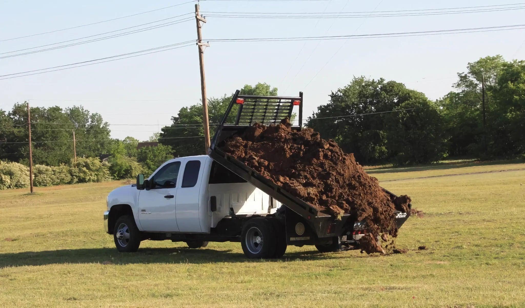 A white dump truck with a PIERCE Medium Duty Flat Bed Dump Kit unloads dirt onto a grassy field under a clear sky.