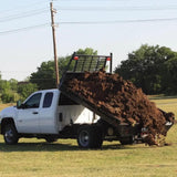 A white truck equipped with the PIERCE Light Duty Flat Bed Dump Kit unloads dirt onto a grassy field under a clear sky.