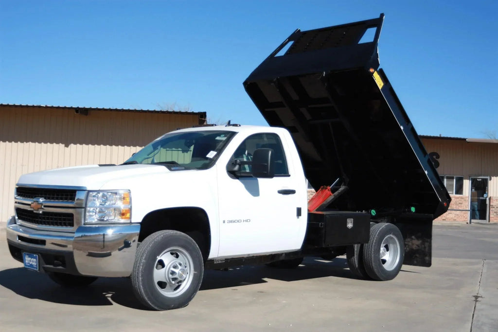 A white truck with a raised black bed showcases the PIERCE Medium Duty Flat Bed Dump Kit, parked outside a tan industrial building beneath a blue sky.