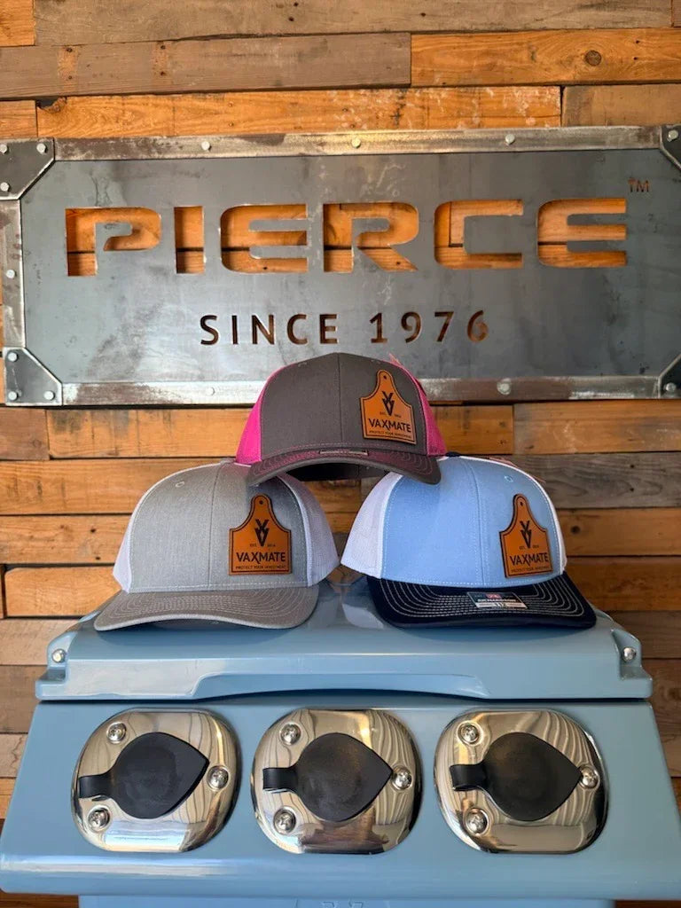 Three VAXMATE Trucker Hats with Leather Patch by PIERCE, in gray, pink, and blue, are shown in front of a PIERCE sign on a wood wall.