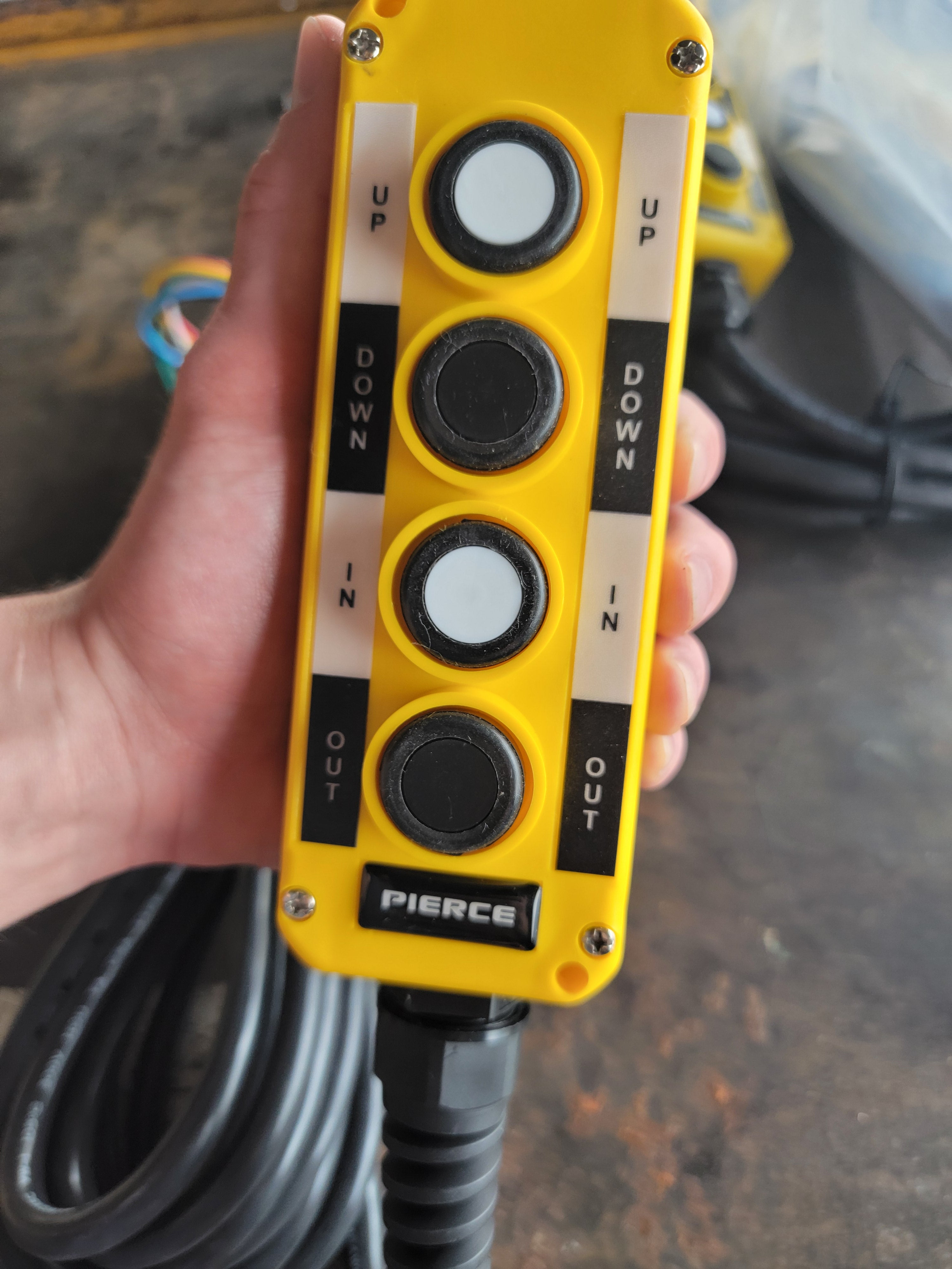 A hand is holding a yellow PIERCE Universal Momentary Pendant Control featuring four buttons labeled Up, Down, In, and Out.