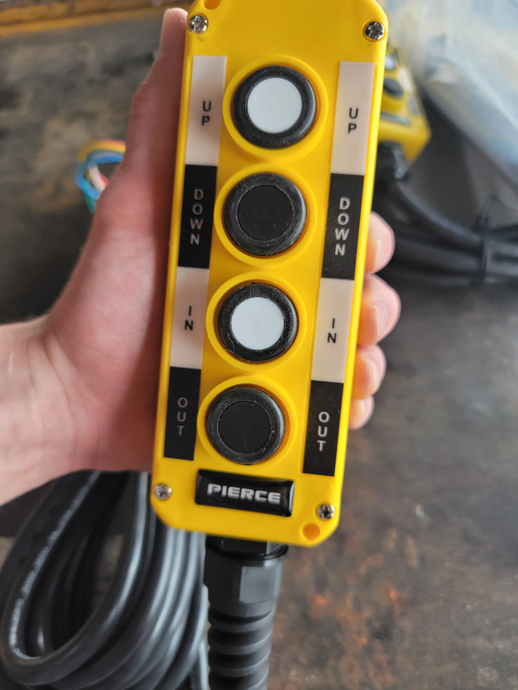 A hand is holding a yellow PIERCE Universal Momentary Pendant Control featuring four buttons labeled Up, Down, In, and Out.