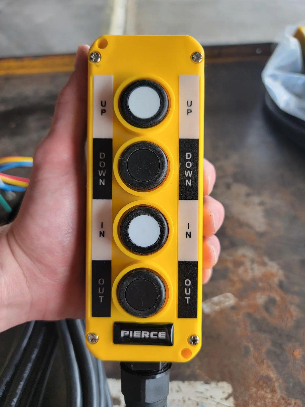 A hand holds the PIERCE Universal Momentary Pendant Control, featuring four labeled buttons (UP, DOWN, IN, OUT), a 20ft cord, and magnets—designed for solenoid-operated equipment.