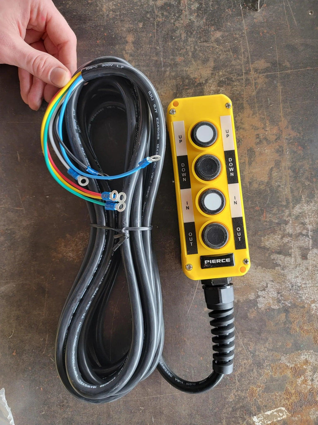 A hand holds the PIERCE Universal Momentary Pendant Control, a yellow 4-button device with a 20ft coiled black cord, magnets, and exposed wires, commonly used to operate solenoid equipment.
