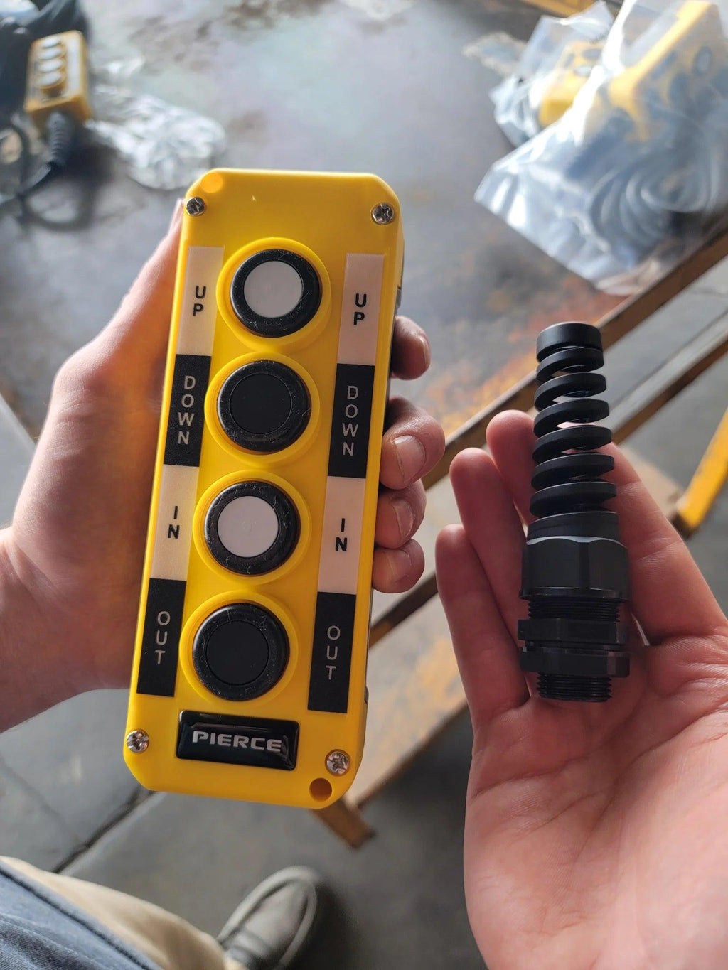 A hand holds the Asetek Enterprise Company LTD Universal Momentary Pendant Control - 4 Button (yellow, with UP/DOWN and IN/OUT), ideal for hydraulic power packs, while the other hand grips a black spring part.