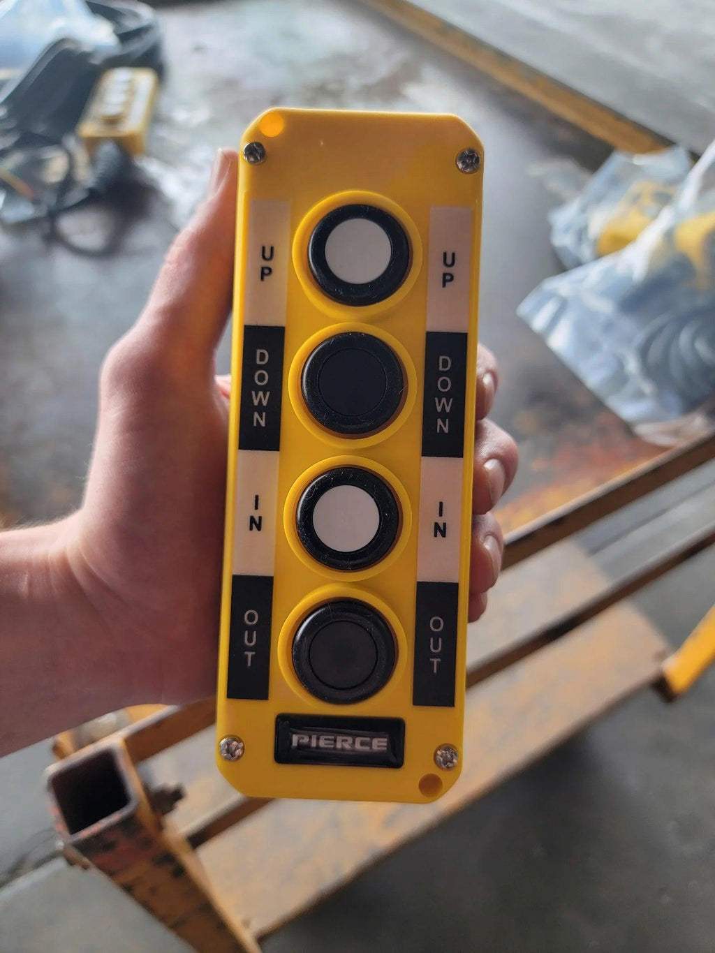 A hand holds the Asetek Enterprise Company LTD Universal Momentary Pendant Control - 4 Button, featuring yellow design and labeled Up, Down, In, and Out—perfect for industrial tasks or hydraulic power packs.