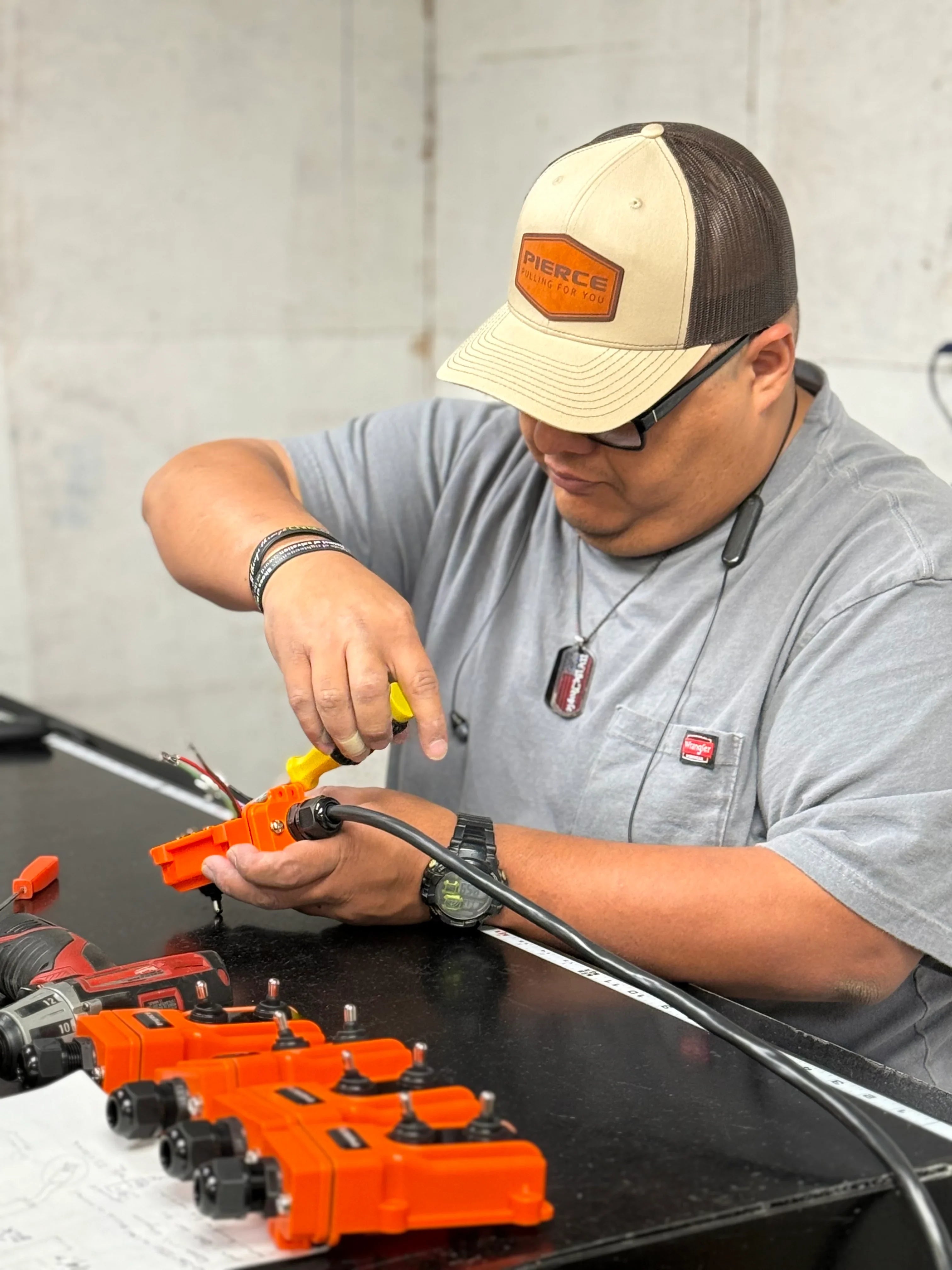 A man in a cap uses a tool to work on orange electrical parts, including a PIERCE 2 function Momentary Hard-Wired Control Pendant with 15 ft. cord and 4 prong vulcanized plug, at a workbench.
