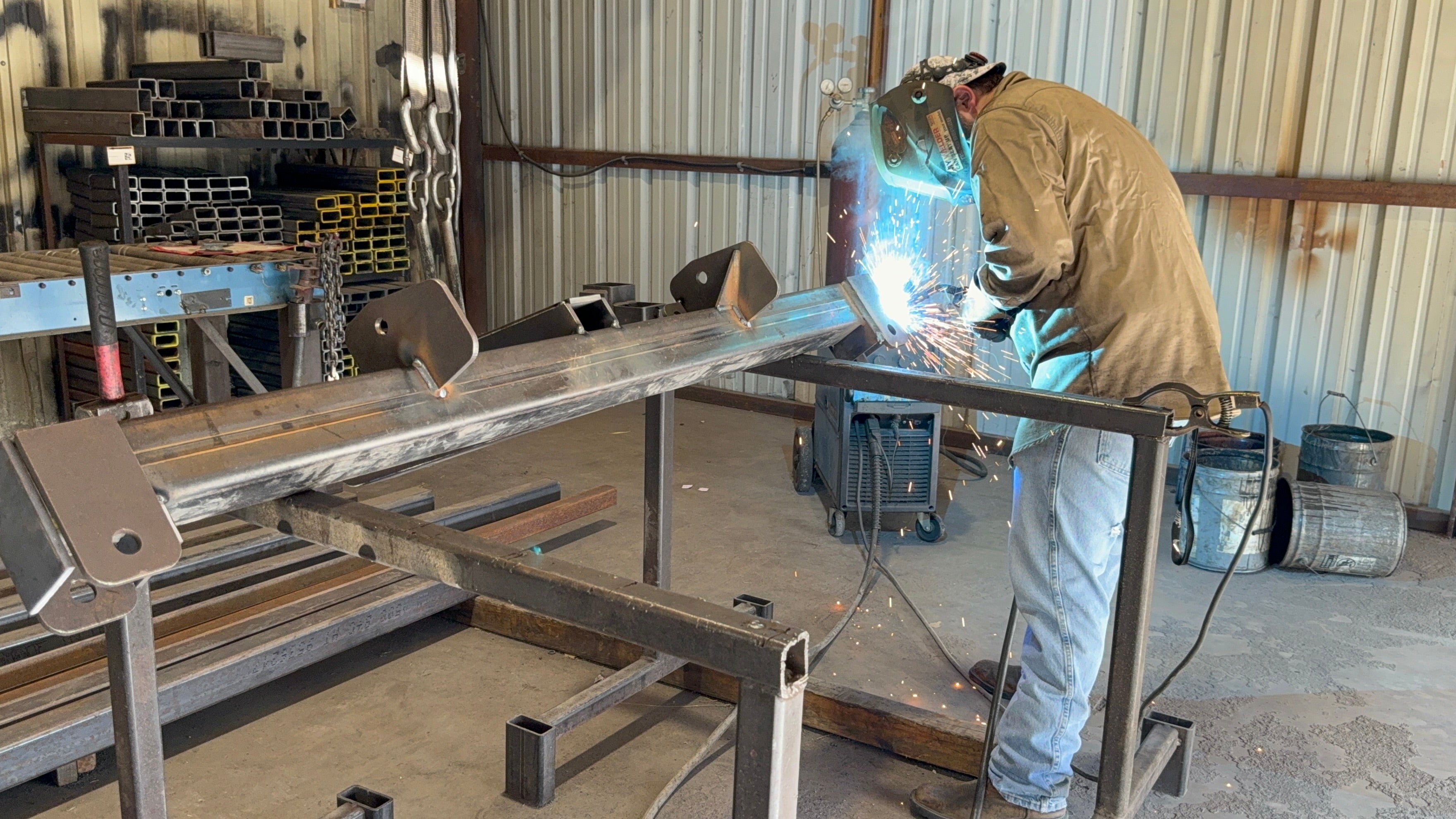 Wearing protective gear, a person welds metal beams in a workshop as sparks fly, fabricating parts for the Internal - Pierce Assembly Team 3-Point Hay Bale Unroller, an agricultural tractor attachment.