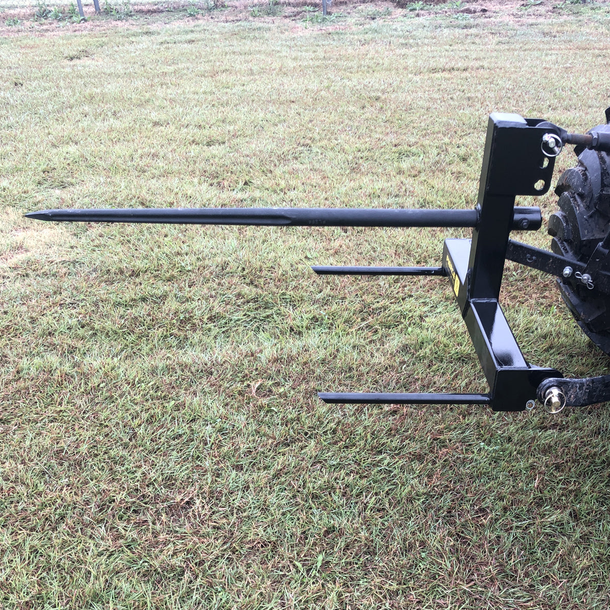 On a grassy field, a black PIERCE 3 Point Tractor Bale Spike made of high-quality steel is attached to the back of a tractor, ready to effortlessly handle round bales.