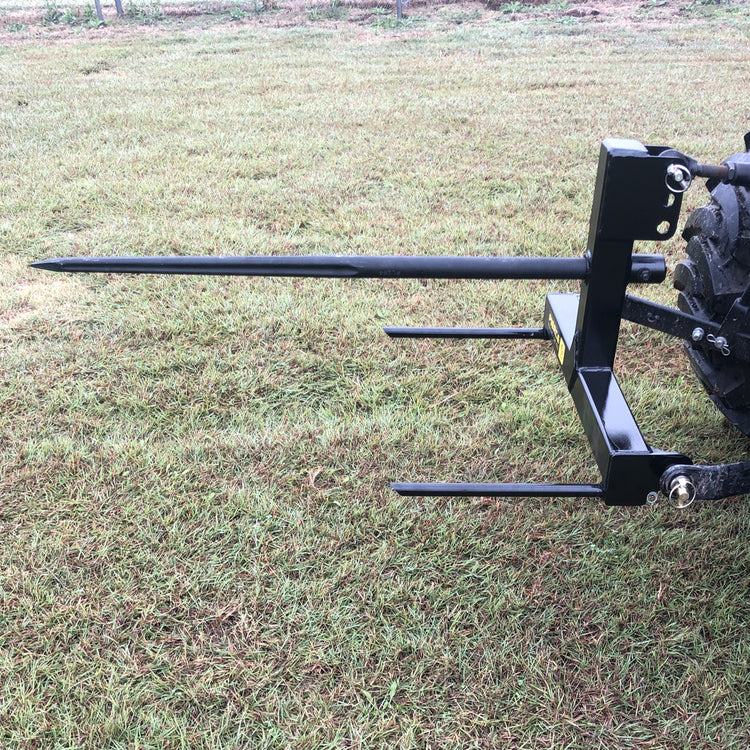 On a grassy field, a black PIERCE 3 Point Tractor Bale Spike made of high-quality steel is attached to the back of a tractor, ready to effortlessly handle round bales.