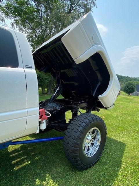 A white truck equipped with a PIERCE Factory Bed Dump Kit displays its lifted suspension and hydraulic dump bed while parked on grass beneath a sunny sky.