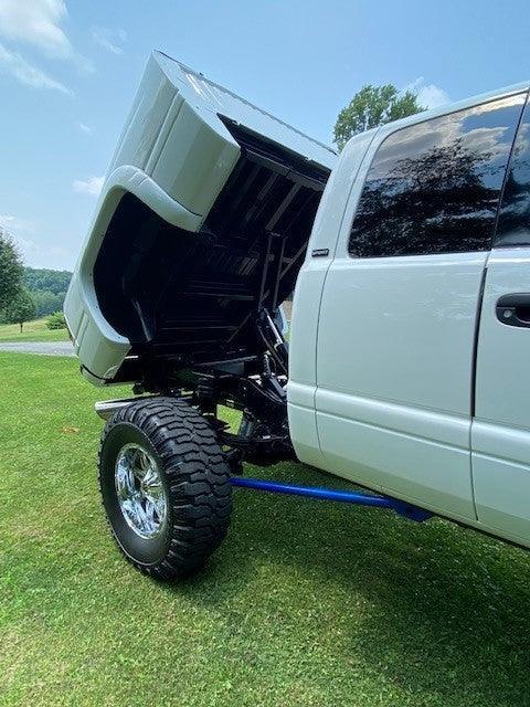 A lifted white truck with its hood up is parked on grass under a blue sky, equipped with a PIERCE Factory Bed Dump Kit.