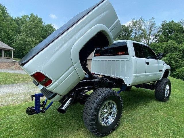 A white lifted pickup truck equipped with a PIERCE Factory Bed Dump Kit, its bed tilted up, is parked on grass near trees.