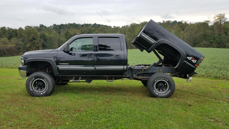 A black pickup truck with a detached, raised bed featuring a PIERCE Factory Bed Dump Kit is parked in a grassy field.