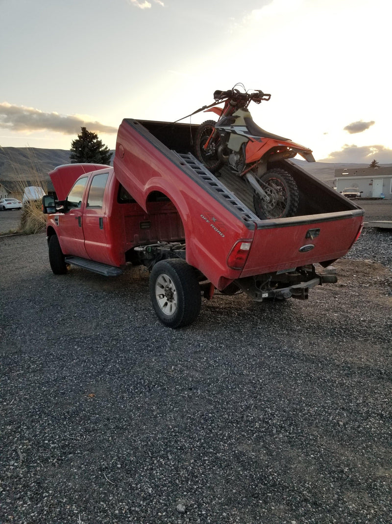 A red pickup truck with a PIERCE Factory Bed Dump Kit tilted upward and a dirt bike in the bed, parked on gravel.
