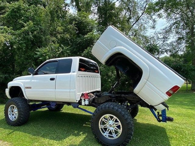 A white lifted truck equipped with a PIERCE Factory Bed Dump Kit is parked on grass, its bed tilted upright with trees in the background.