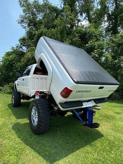 A white Dodge truck equipped with a PIERCE Factory Bed Dump Kit has its bed tilted up, parked on grass with trees in the background.